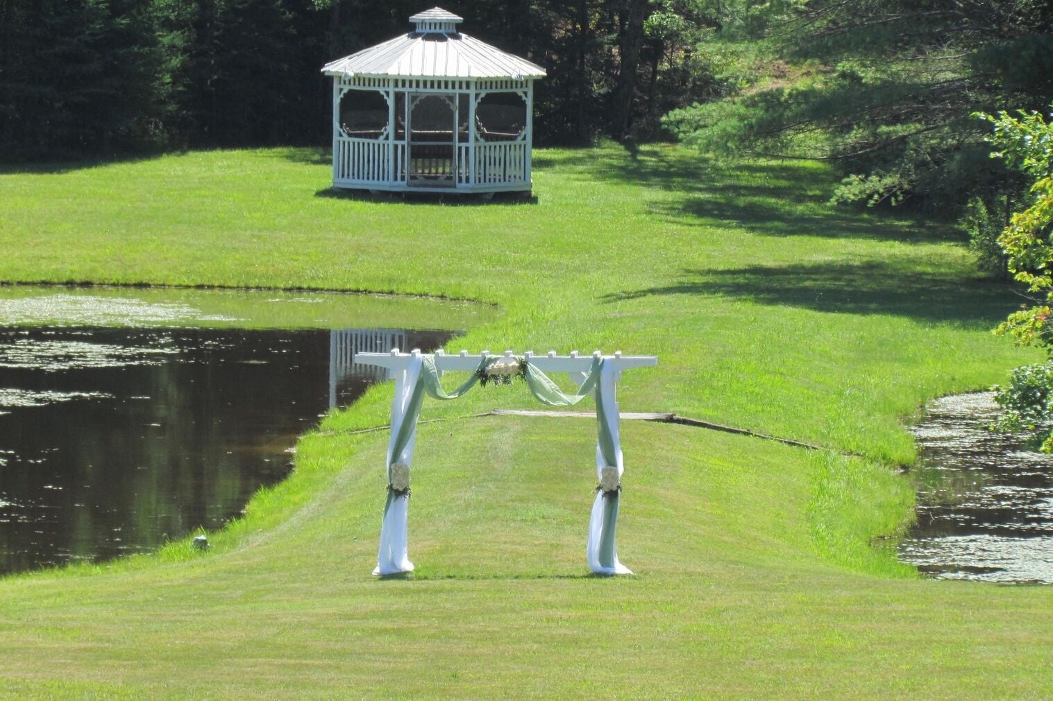 A white gazebo sits near a pond in a grassy area, with a decorated wedding arch in the foreground on a mowed lawn.