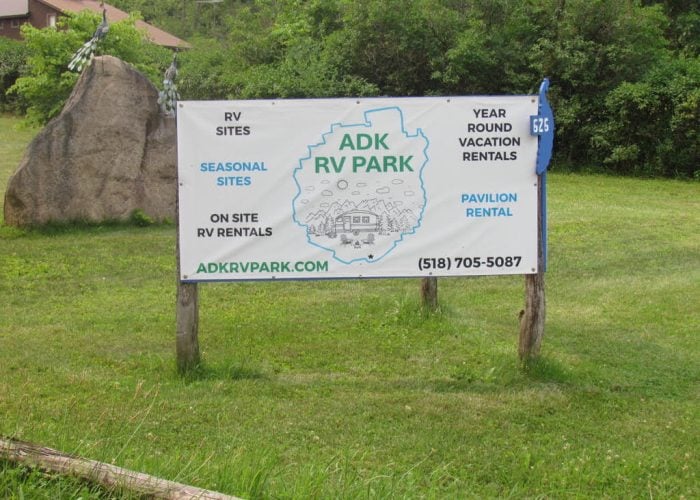 ADK RV Park sign on grassy area shows rental info, website, and phone number. Services include RV and vacation rentals, seasonal and onsite options, and pavilion rental.