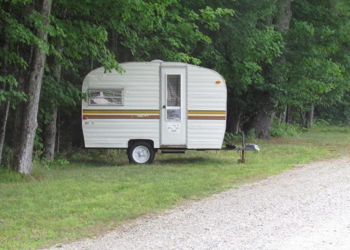 A small vintage camper trailer with a brown stripe is parked on grass near trees, beside a gravel road.