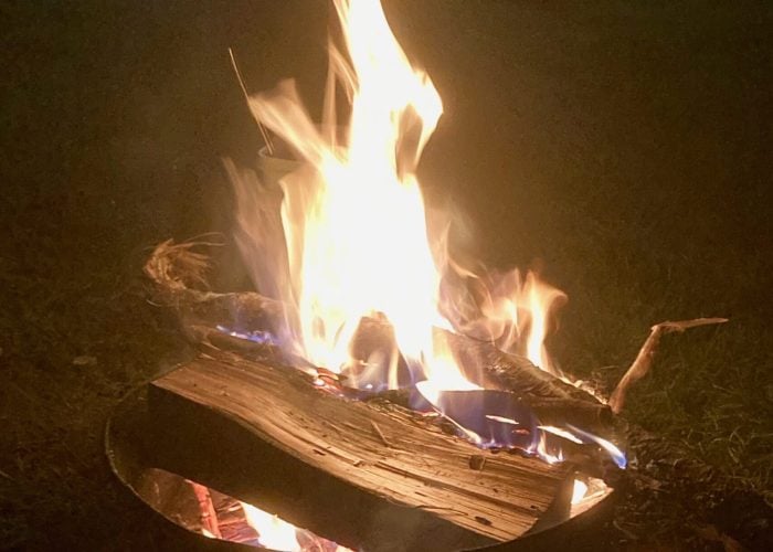 A campfire burns brightly in a metal fire pit at night, with wooden logs and visible blue and orange flames. Outdoor furniture and steps are visible in the background.