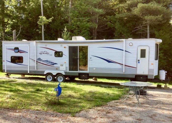 A white and silver travel trailer with blue and red stripes is parked on grass near trees. A round glass table and chairs sit on gravel in the foreground.