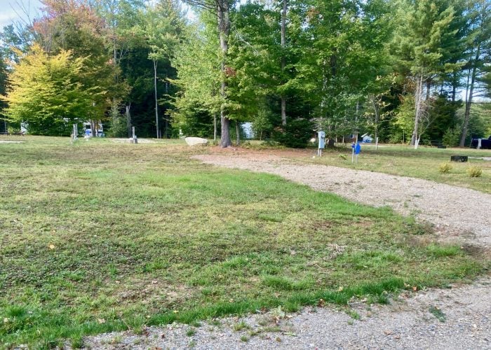 A gravel path curves through a grassy area with scattered trees, utility posts, and parked vehicles in the background.