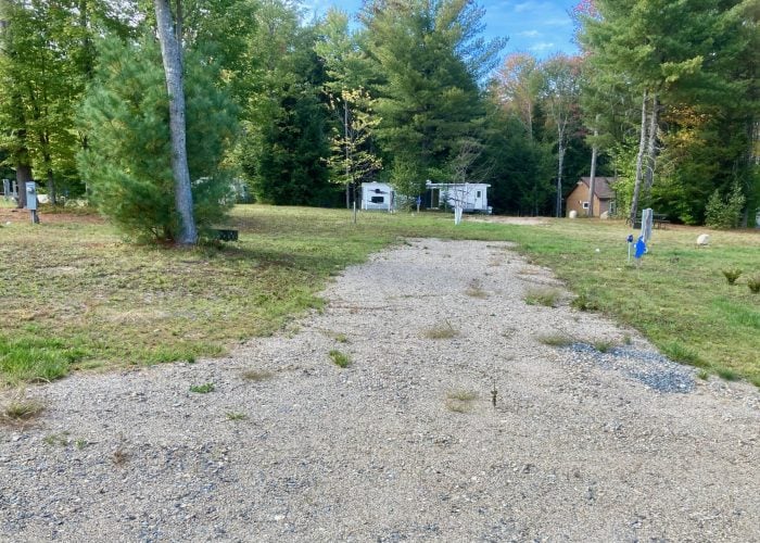 A gravel campsite with electrical and water hookups, surrounded by grass and trees, with RVs and a cabin visible in the background.