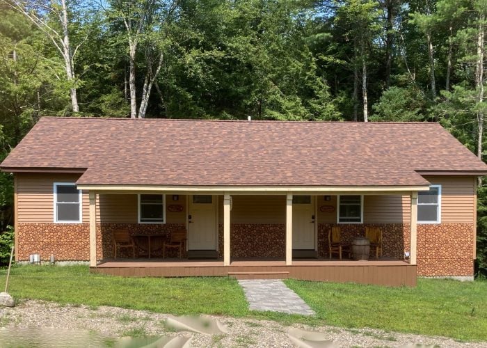 A tan and brown duplex cabin with a covered porch, two doors, outdoor chairs, and surrounded by green trees and grass.