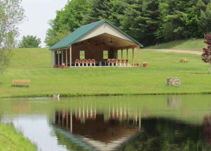 A pavilion with a green roof sits on a grassy hill near a pond, surrounded by trees. A bench and a tree stump are visible by the pond’s edge. The pavilion is reflected in the water.