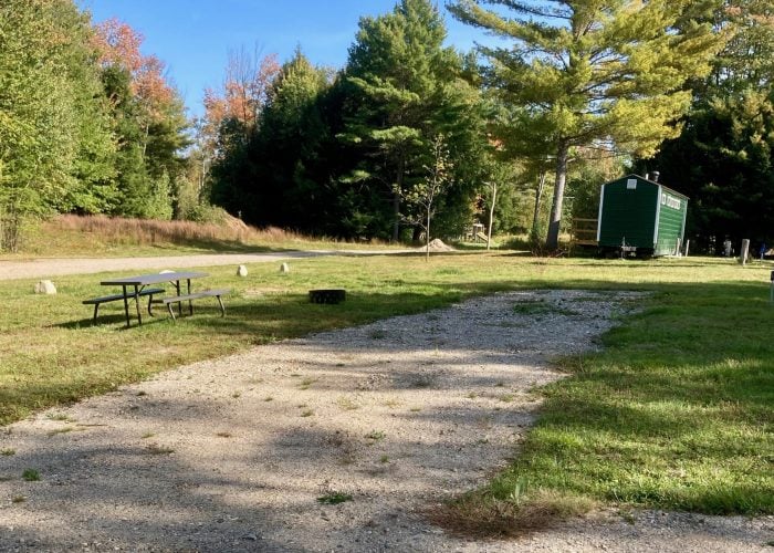 A gravel driveway leads to a grassy area with a picnic table, fire pit, and a small green cabin surrounded by trees under a clear blue sky.