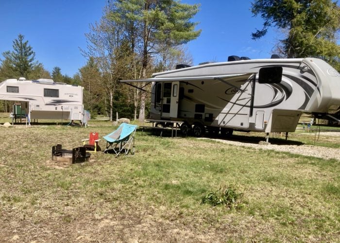 Two RVs are parked on a grassy campsite with trees in the background. In front, there is a fire pit, a folding chair, and a bucket. The sky is clear and blue.